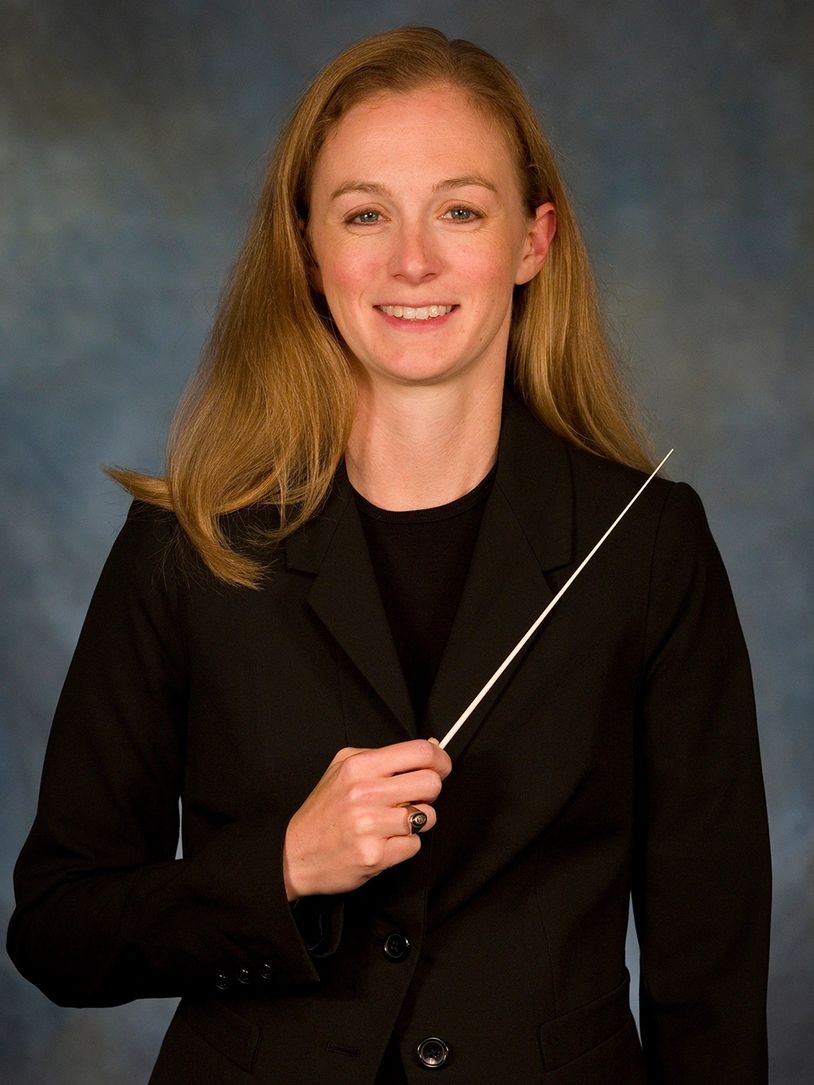 Woman in black suit holding a conductor's baton, smiling.
