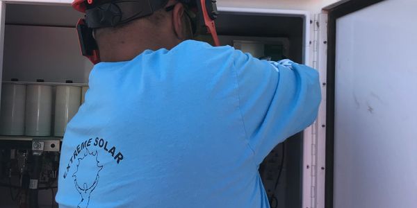 Worker in a blue shirt and red helmet fixing electrical equipment under solar panels.