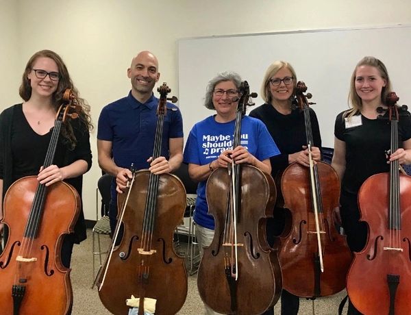 Five smiling musicians holding cellos in a room with a whiteboard.