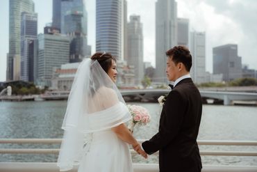 Bride and groom holding hands by the waterfront with city skyline behind.
