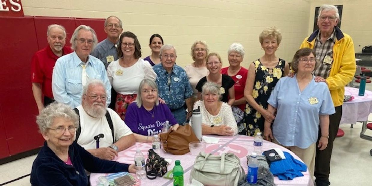 A group of older adults gathered indoors around tables, smiling for a photo.