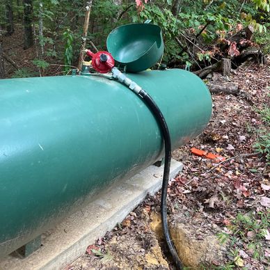 Large green propane tank with hose connected, outdoors in wooded area.