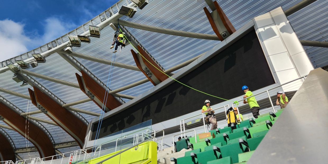 Workers perform high-altitude maintenance on a stadium roof with safety harnesses.