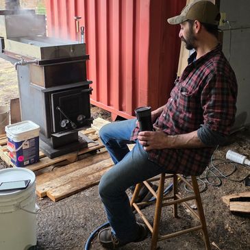 Man sitting on a stool next to a steaming metal stove.