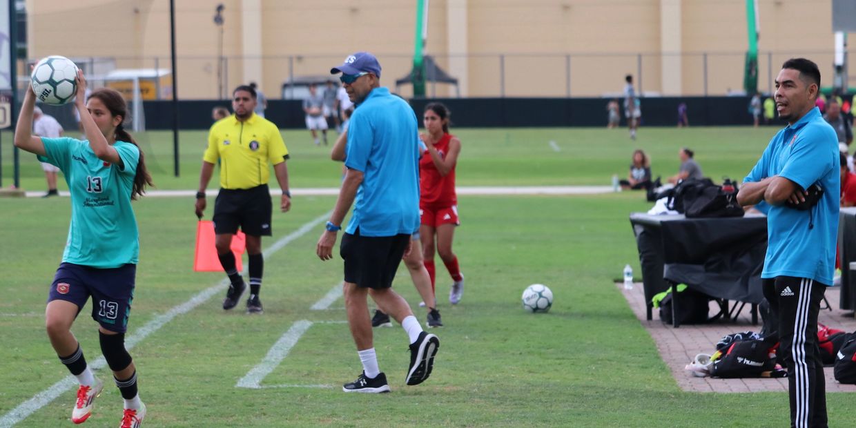 A youth soccer player prepares for a throw-in with coaches and referee nearby.