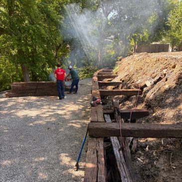 Two men working on a wooden retaining wall with smoke rising nearby.