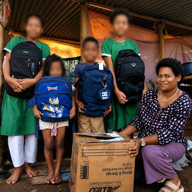 Group of children and a woman with backpacks and a cardboard box outside a rustic building.