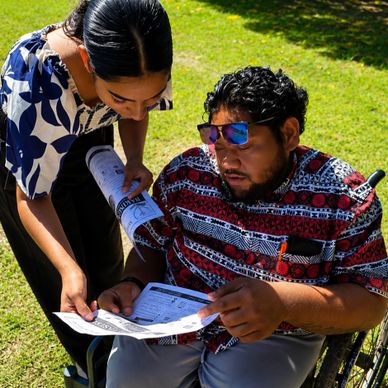 A man in a wheelchair and a woman review documents outdoors on a sunny day.