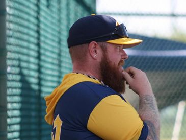 A bearded man in a baseball cap and jersey watches intently.