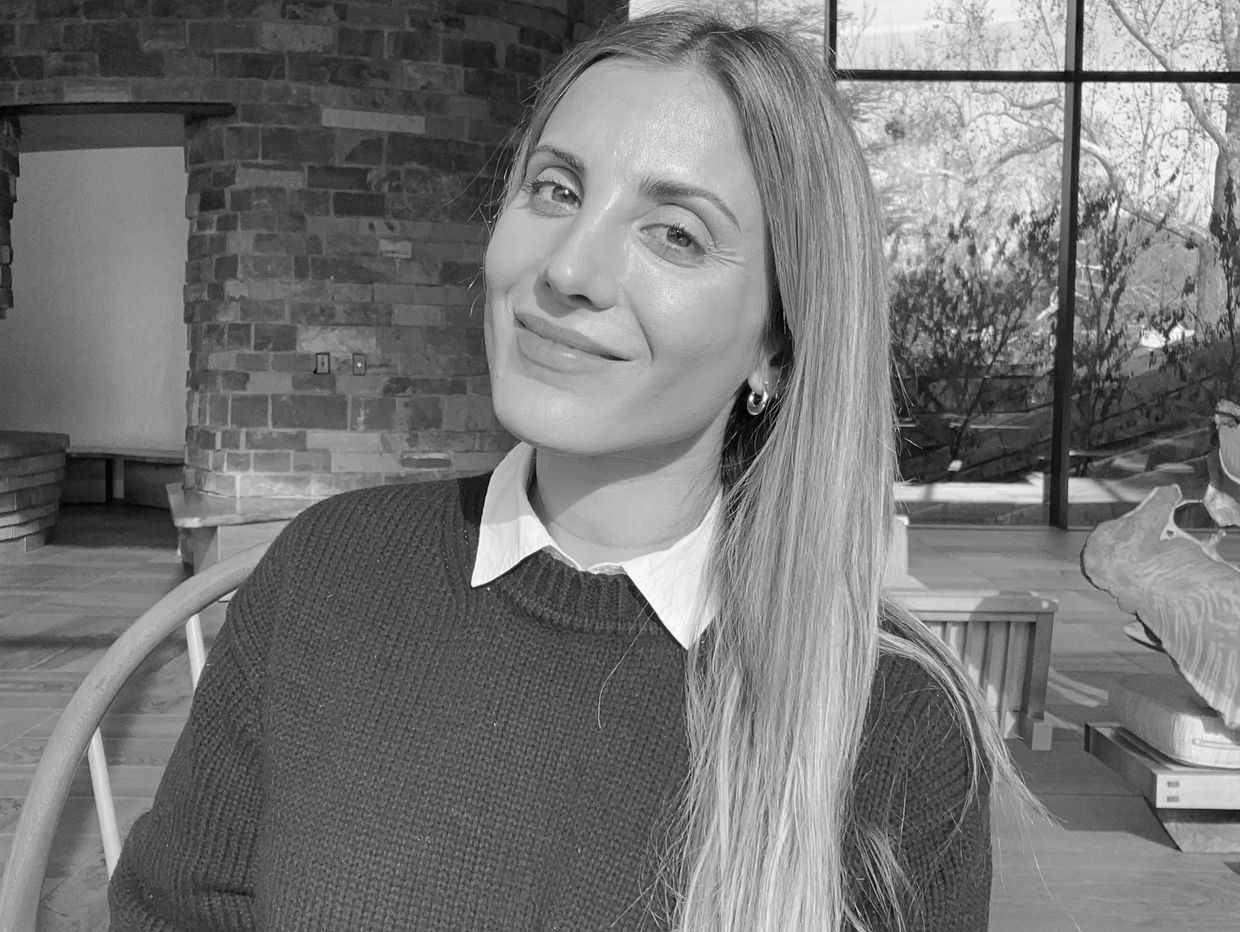 Smiling woman in a cozy maroon sweater sitting indoors by a stone wall and window.