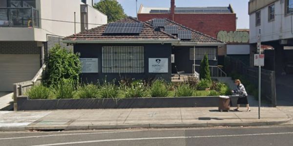 Small dark building with solar panels and greenery in front on a city street.