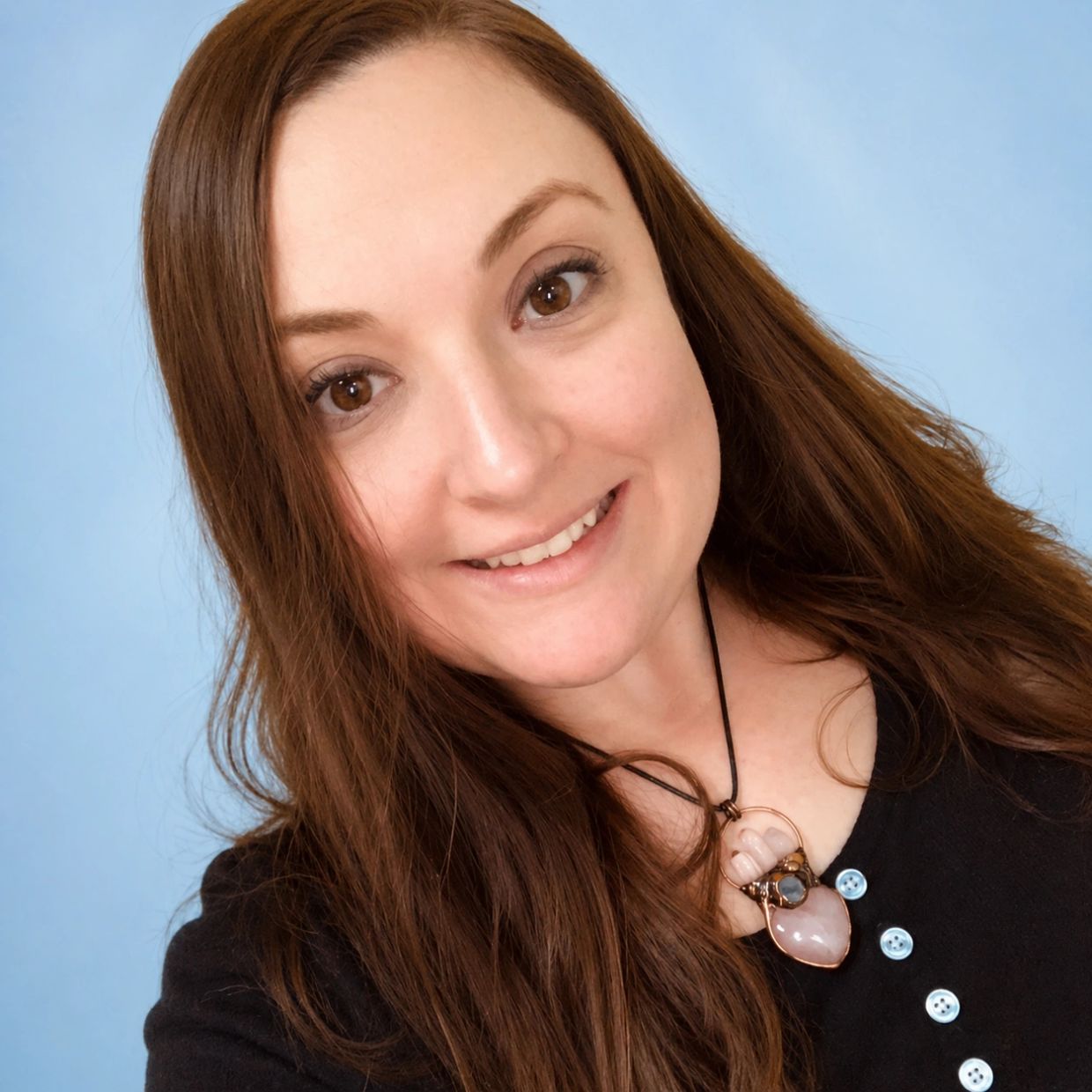 Smiling woman with long brown hair wearing a black top and a gemstone necklace.