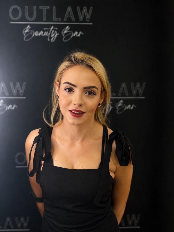 Young woman posing in front of Outlaw Beauty Bar sign, wearing black dress.