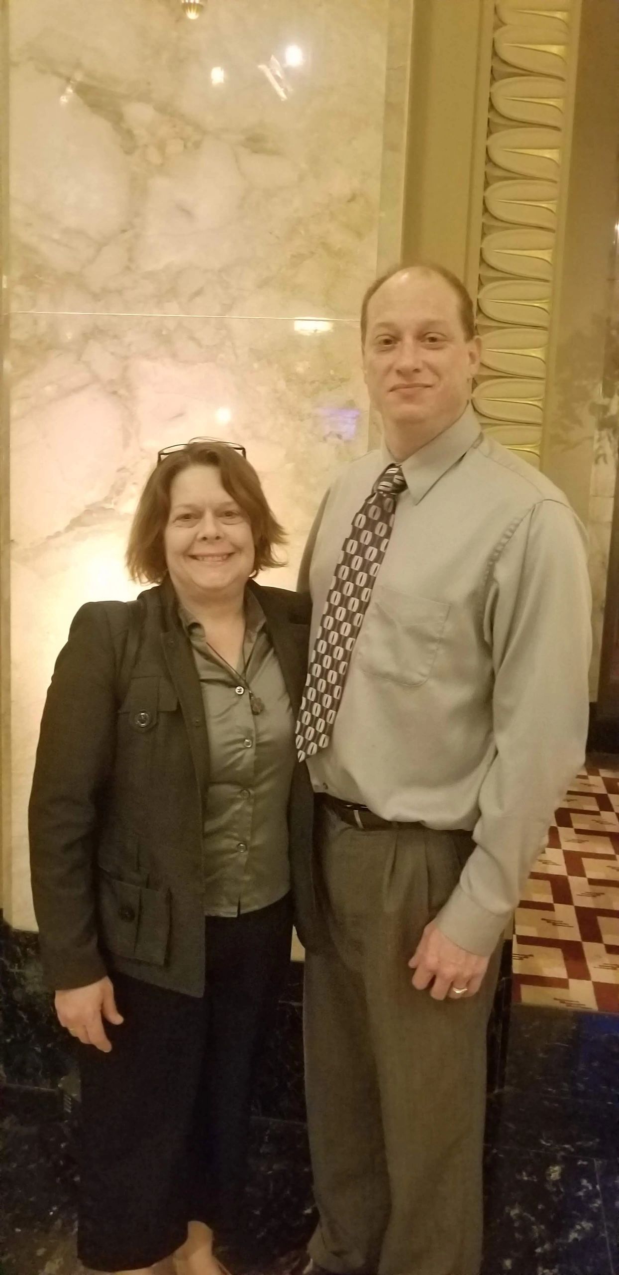 A smiling couple standing indoors against a marble wall.