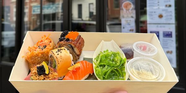 Hand holding a sushi box with rolls and seaweed salad in front of All Buru restaurant.