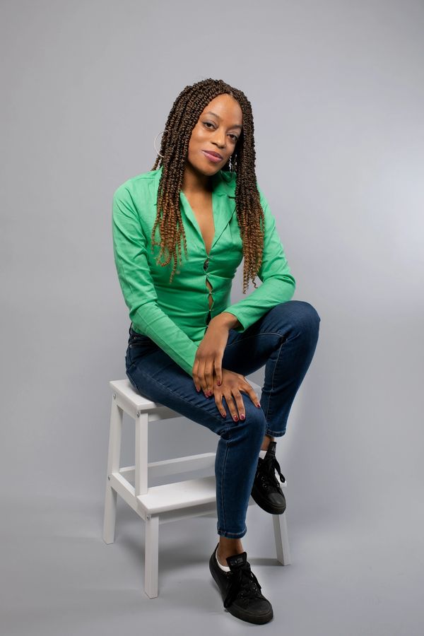 Woman with braided hair wearing a green blouse and jeans, sitting on a white stool.