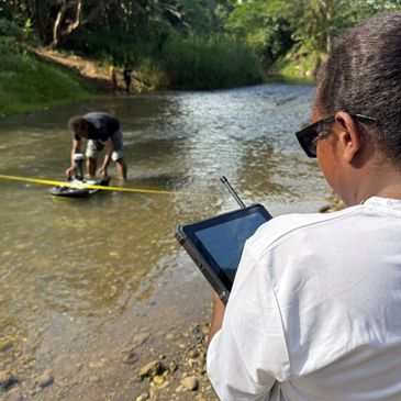 Person using a tablet while another measures water in a river.