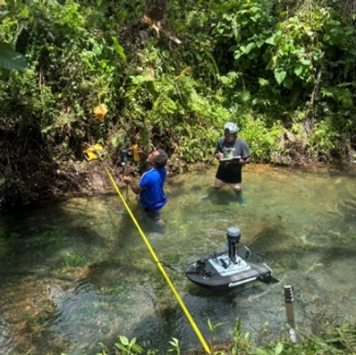 Two people working with a remote-controlled boat in a shallow stream surrounded by greenery.