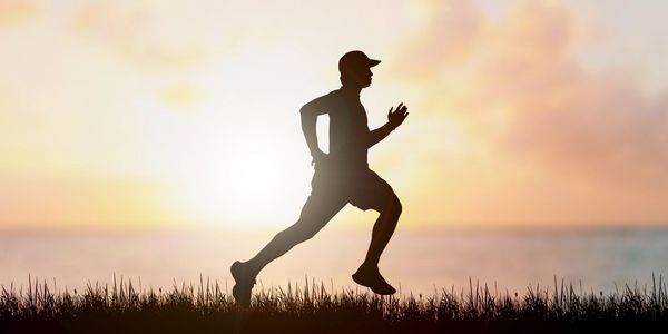 Silhouette of jogger against sunrise.  Jogger is running through grass beside a lake.