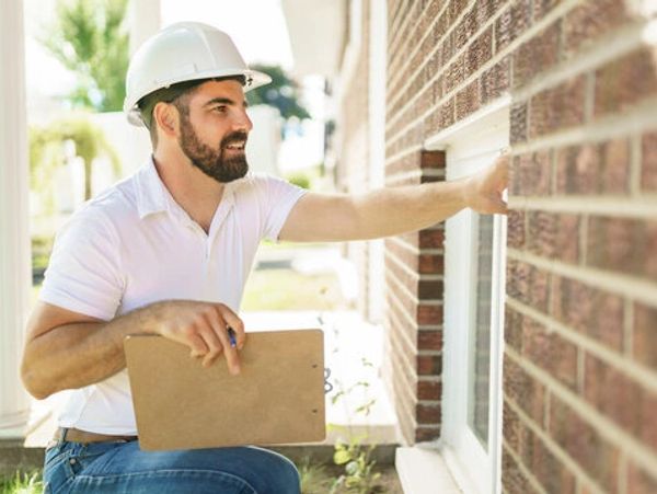 Man in a hard hat inspecting a house exterior with clipboard.