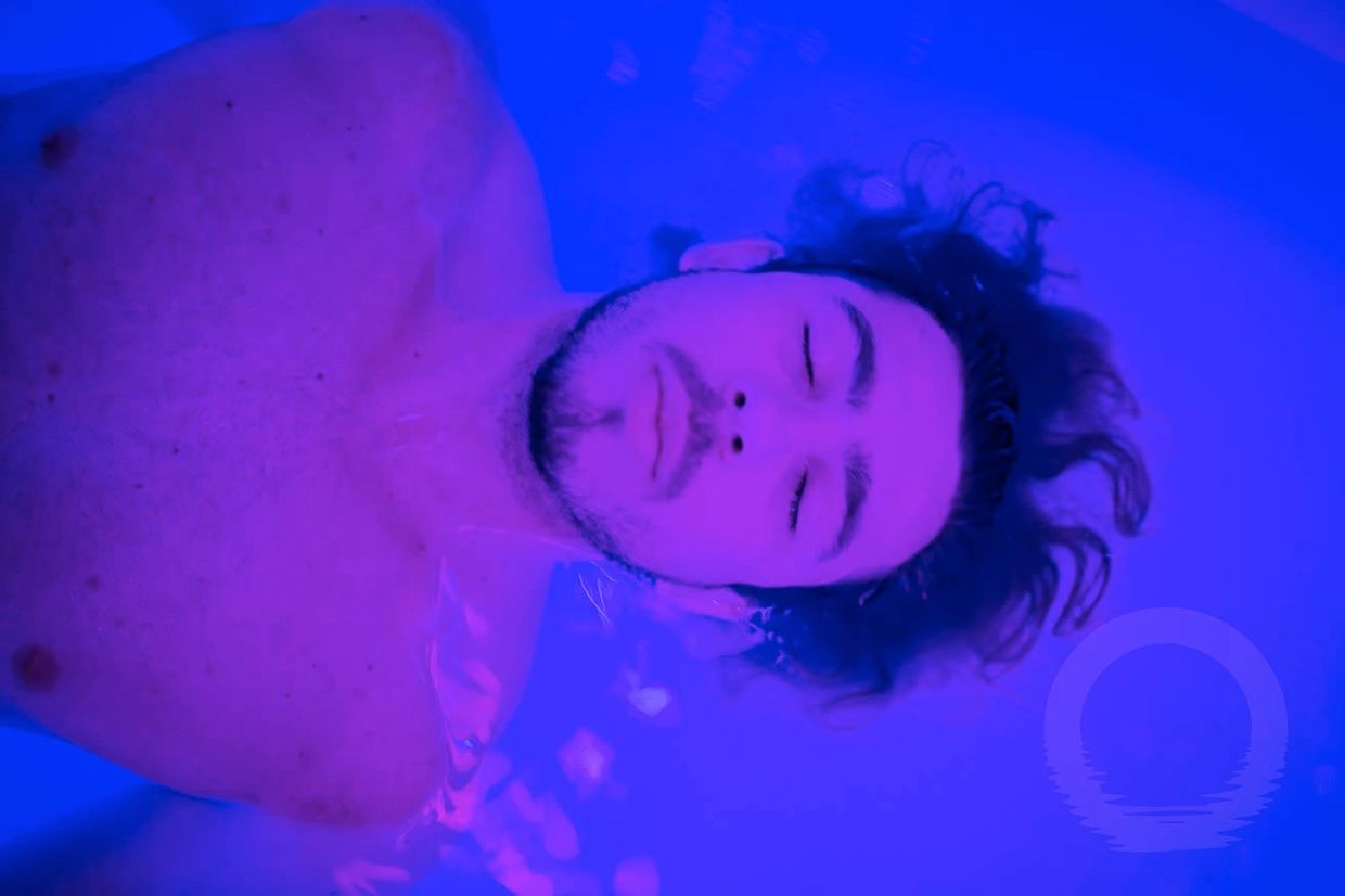 Man relaxing in water under blue lighting with eyes closed.