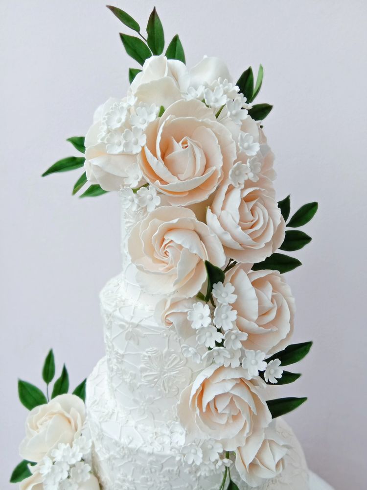 Large ivory hand-sculpted sugar roses cascading down a white lace fondant cake, accented with delicate white blossoms and deep green ruscus leaves. This sophisticated gum paste floral arrangement is a professional-grade wedding cake topper, ideal for luxury celebrations and classic-meets-modern bridal designs. Artisan sugarcraft by Bespoke Sugarcraft.