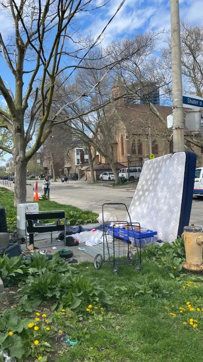 Discarded mattress and household items on a sidewalk near a tree and street.