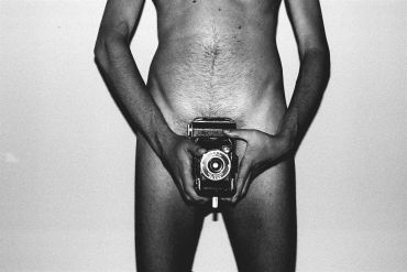 Homem nu segurando uma câmera vintage na frente do corpo.