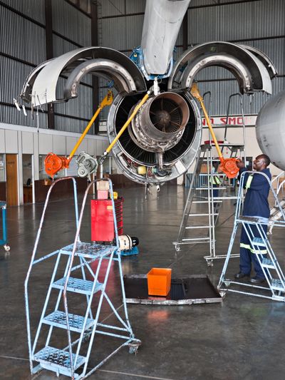 Man works on a large jet engine with its cowling open, surrounded by tools and ladders in a hangar.