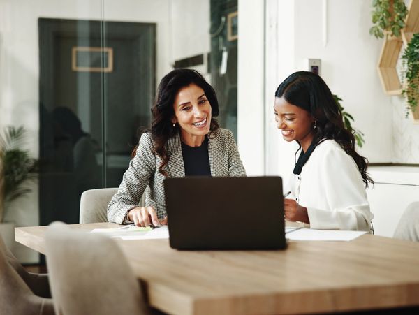 Two smiling women, one looking at a laptop and the other writing, collaborate at a conference table.