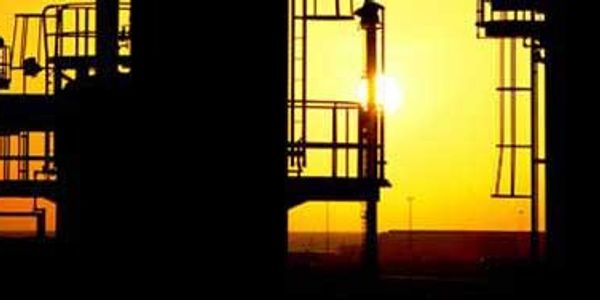 Industrial structures are silhouetted against a vibrant orange sunset, with a sunburst visible.