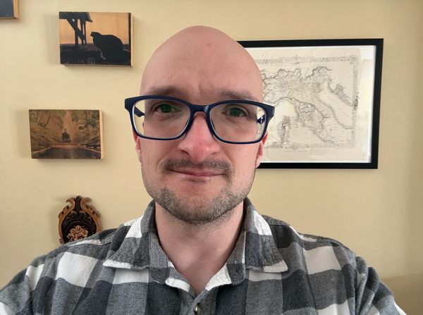 Man with glasses and a plaid shirt smiling indoors with framed pictures in background.