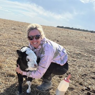 A smiling woman kneels in a field hugging a young calf beside a feeding bottle.