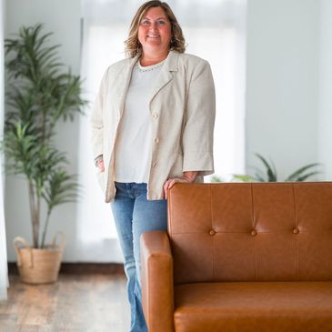 Smiling woman standing beside a brown leather sofa in a bright room.