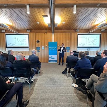 A speaker presents to a seated audience in a professional conference room.