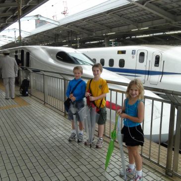 Three children standing with umbrellas near a bullet train at a station.