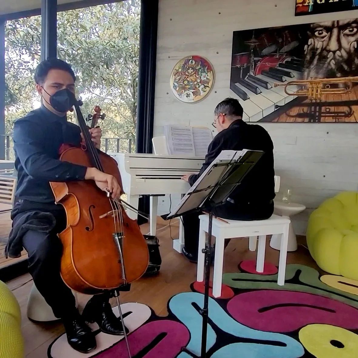 Two musicians playing cello and piano in a cozy room with colorful decor.