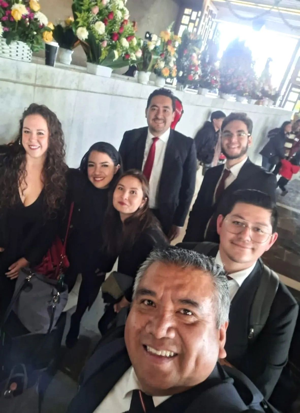 Group of smiling people posing for a cheerful selfie indoors with floral arrangements in the background.