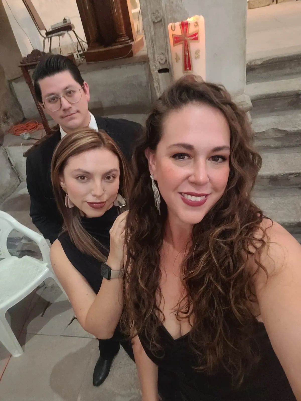 Three well-dressed friends smiling for a selfie inside a church with a lit candle.