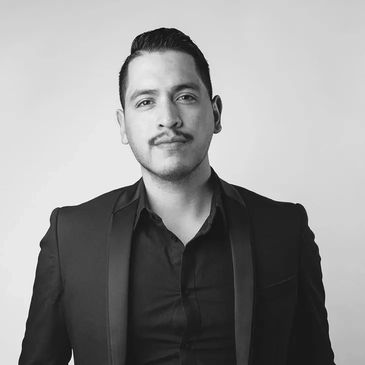 Man in a black suit posing confidently against a plain background.