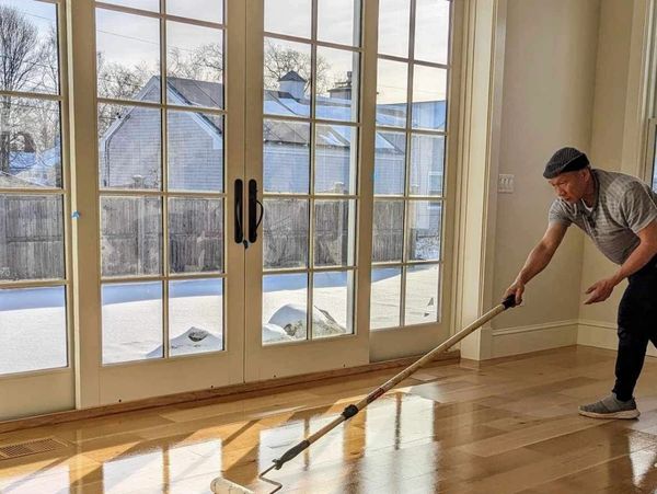Man applying finish on wooden floor with roller in sunlit room.