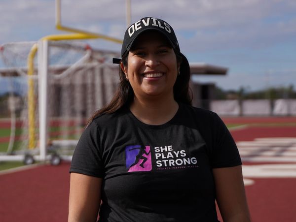 Smiling woman wearing a 'Devils' cap and 'She Plays Strong' t-shirt on a sports field.