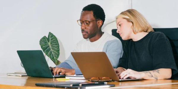 Two colleagues collaborate on laptops at a wooden table in a modern office.