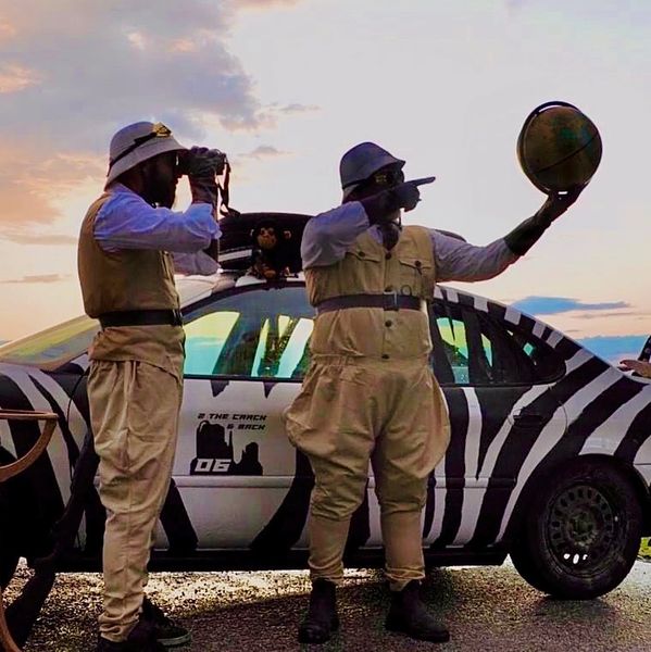 Neil and Daryl, dressed up in Safari attire, pointing and looking at a globe through binoculars. The