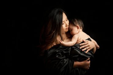 A mother tenderly holds her baby wrapped in black fabric against a dark background.