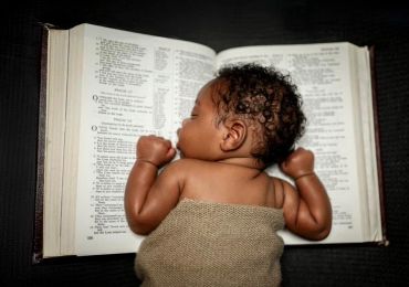 A baby sleeps on an open Bible, wrapped in a cloth.