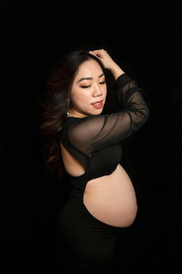 Pregnant woman posing gracefully in a black dress against a dark background.