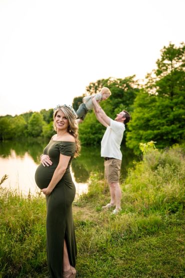 Pregnant woman smiles by a lake while man lifts a child in the background.