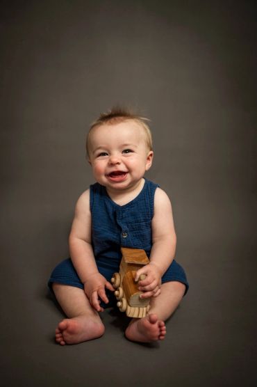 Smiling baby in blue romper holding a wooden toy, sitting against a gray background.