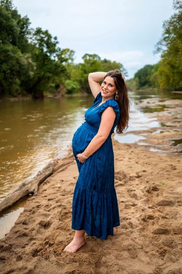 Pregnant woman in a blue dress poses barefoot by a river on sandy shore.